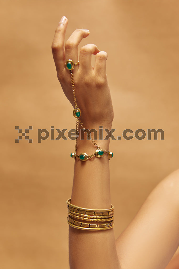 Elegant hand adorned with gold and emerald jewelry, featuring a chain bracelet and stacked bangles against a warm-toned background stock image.