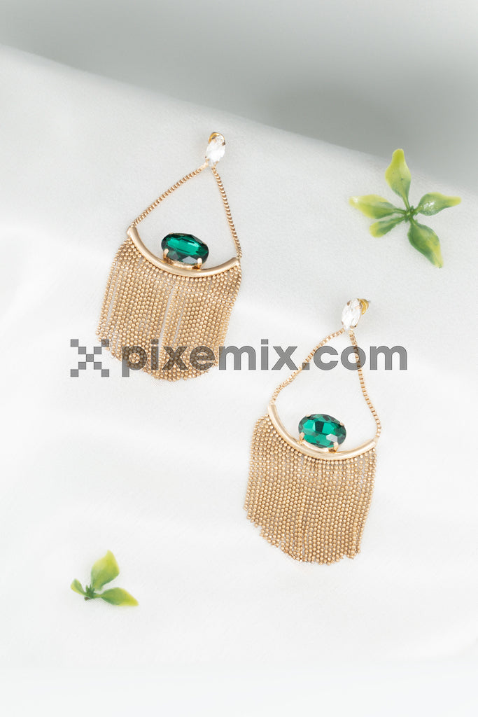 Elegant gold fringe earrings with emerald gemstones displayed on a soft white fabric background stock image.