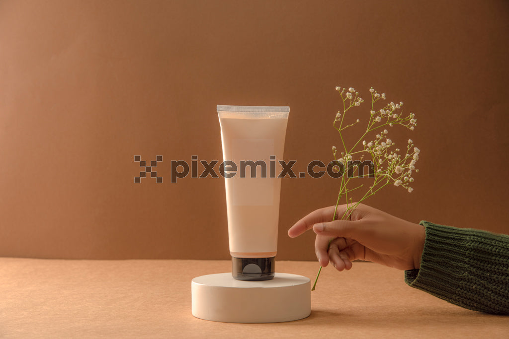 Hand holding flowers next to a skincare product tube on pedestal with warm brown background stock image.