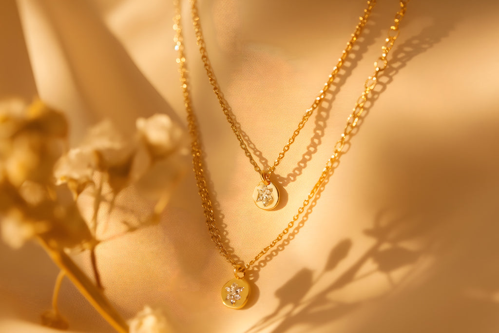Elegant gold layered necklaces with delicate pendants captured in soft golden light and floral shadow.