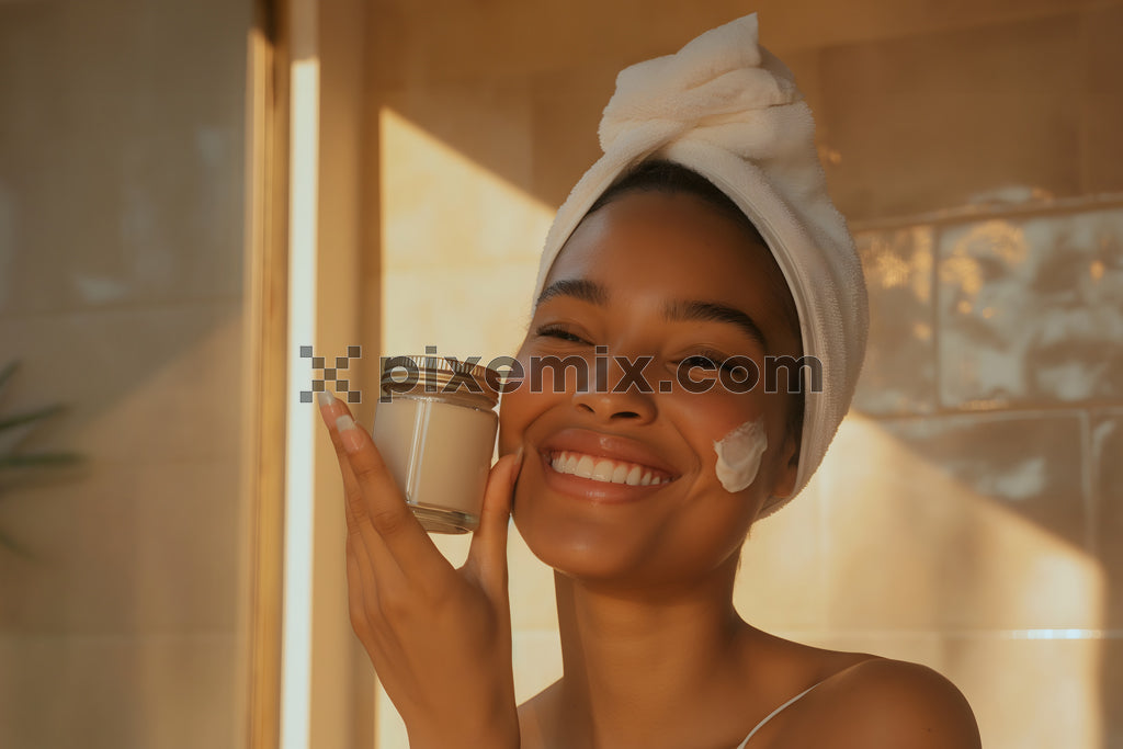 Radiant woman with towel on head applying face cream and smiling in warm morning light.