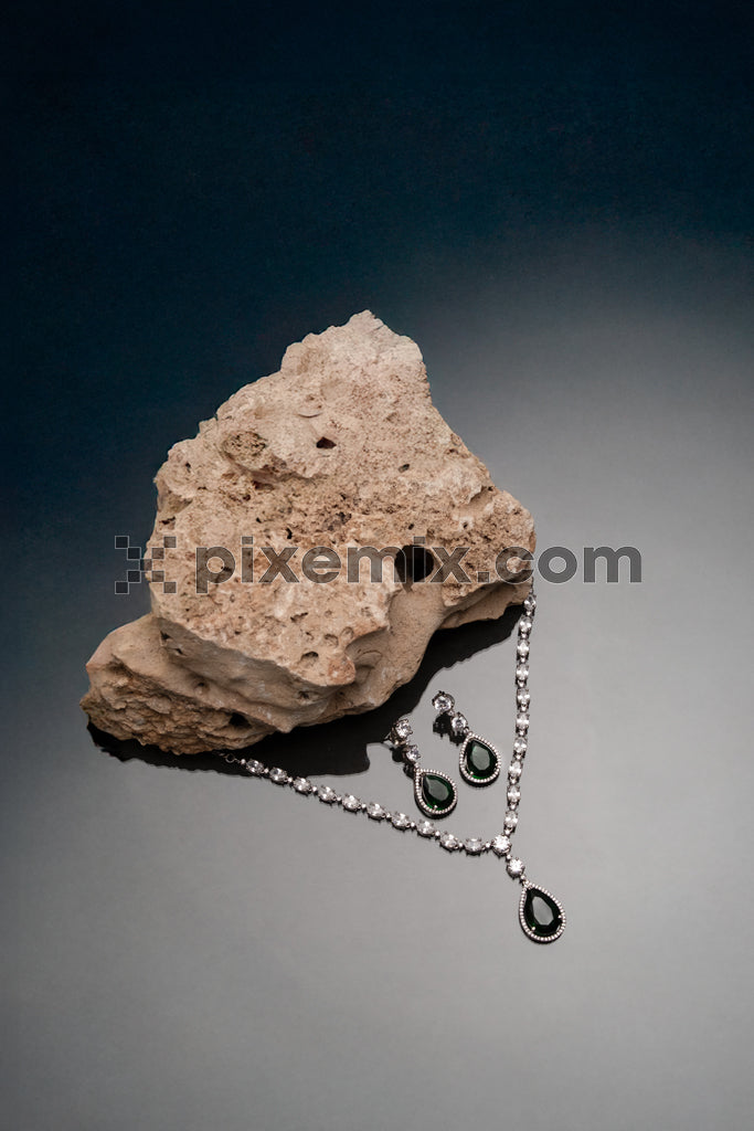 Elegant emerald and diamond jewelry set displayed on stone with dark moody gradient background.