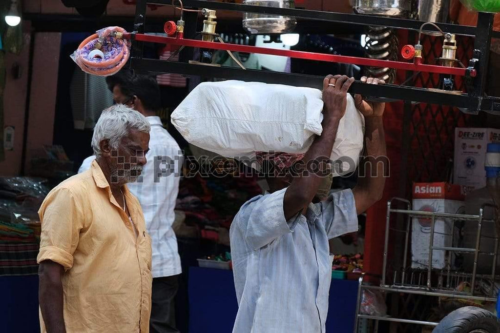 Two old indian men walkng in streets with one carrying heavy luggage on head