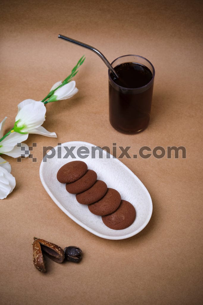 A plate of chocolate cookies and a glass of black coffee with a metal straw sit on a light brown background with white flowers image.